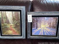 Two framed oil paintings, one of forest path and one snowy forest, displayed on dark couch.