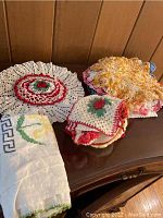 Photo showing multiple crocheted doilies with floral patterns in red, green, yellow, and white, along with folded cloth napkins on wooden surface.