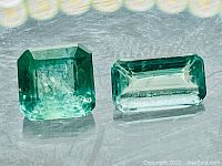 Two emerald gemstones side by side on reflective surface showing color and cut