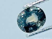 Close-up of oval cut sapphire held by metal tweezers, displaying green hue.