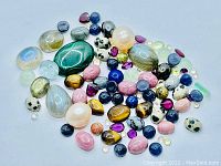 Photo shows a collection of various polished and bead-shaped natural gemstones including Labradorite, Tigers Eye, Dalmatian Jasper, Emerald, Blue Jade, Pearls, and others in multiple colors and sizes.