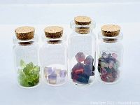 Front view of four small glass bottles each containing a different type of gemstone: green Peridot, purple and yellow Ametrine, red Garnet, and multicolour Tourmaline, all with cork stoppers.