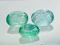 Three light to medium green natural emeralds of mixed sizes shown on a white reflective surface, highlighting the oval faceted shape and internal characteristics.