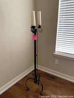 Photo showing a black wrought iron candle holder with two white taper candles, placed against a wall on wooden floor.