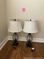 Two identical table lamps with white shades and decorative glass and metal bases, placed on wooden floor against cream wall.