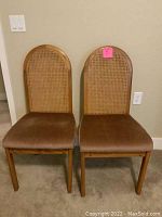 Two vintage wooden chairs with cane woven backs and brown cushioned seats against a wall on carpeted floor.