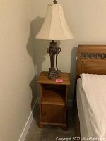Small wooden side table with single door cabinet and open shelf, topped with vintage style lamp base and cream fabric shade next to bed.