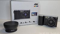 Digital camera with black body alongside the original packaging and a detachable macro lens labeled 'MACRO'. Box shows 4K Full HD Video branding and highlights features such as 24MP resolution and 8x digital zoom.