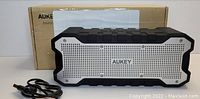 Front view of Aukey Bluetooth speaker in black and silver with USB charging cable in front and original packaging box behind.