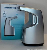 Photo of the automatic soap dispenser showing a sleek silver body with a box in the background highlighting features like adjustable dosage, human ergonomic design, and waterproof rating.