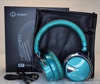 Photo shows Cowin E7 dark green noise canceling headphones, auxiliary and charging cords, carrying pouch, and product box arranged against a neutral background.