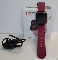 Smart watch displayed vertically in front of white branded box with orange accents. Charging cable shown coiled beside watch.