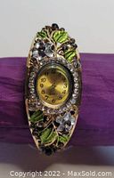 Photo of gold-tone bangle-style watch with green and blue enamel leaves and flowers. Clear rhinestones set around the watch face. The watch face is oval and gold-colored with black numbers and hands.