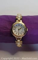 Front view of a gold-tone circular watch with a white dial, black numerals, and a bracelet accented by pearl and crystal details.