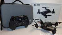 Holy Stone HS165 drone, remote controller, and gray carrying case arranged side by side showing drone unfolded with visible camera lens and controller layout.