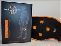 Photo showing Myovolt Knee Kit black wearable knee wrap with orange trim alongside its box packaging with branding and usage visuals.