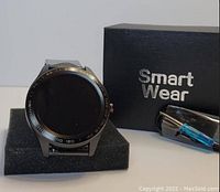 Front image of the black metal Smart Wear smartwatch on a foam cushion, with the Smart Wear branded box and screwdriver accessory beside it.