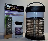 Photo of solar bug zapper lantern next to its retail packaging box. The box highlights features including 360 degree trapping mosquitoes, UV light attraction, and chemical/odor free.