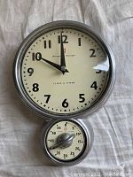 Front view of wall clock showing clock face with black numeral markers and black hands, small timer at the bottom.