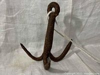 Front view of the small rusty iron dory anchor on a white fabric backdrop with a rope looped through the eye at the top.