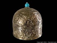 Front view of the vintage metal box showing embossed floral and scroll patterns, decorative clasp, and turquoise finial on the lid.