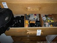 Photo showing a partially open drawer with multiple cameras, camera accessories, and film packs, as well as a black camera bag hanging on the left side.