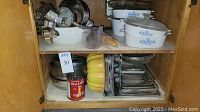 Kitchen cabinet shelves displaying assorted bakeware, casseroles, bowls, rolling pins, and a coffee can. Visible items include metal mixing bowls, yellow plastic bowls, metal cookie cutters, and Pyrex casseroles.