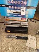 Stack of electronic devices including Sony DVD/VHS player in original blue box, Memorex Power Center box, Philips DVD player, and Sony VHS VCR with two remote controls and user manual on carpet.