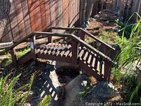 Front angled view of wooden garden bridge spanning a small rock area. Shows brown stained wood with handrails and slatted decking.