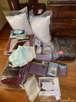 Wide shot showing two new white pillows, various bedding items including sheets, pillowcases, a red velour blanket, and a plastic bag with leopard print bedding.