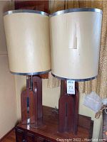 Full view of the pair of Mid-Century Modern wood table lamps with cream cylindrical lampshades trimmed in silver, lampshades show signs of wear and damage.