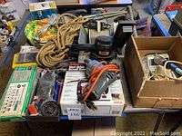 View of table-top showing corded drill with orange cord, Skill Vari-Orbit scroll saw box, miscellaneous tools, rope, and hand tools spread on table.