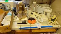 Wide view of table with various small kitchen appliances including the Cuisinart food processor, popcorn maker, hand mixers, cutting tool, Salad Shooter, crock pot