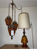 Photo of three amber glass swag pendant lamps hanging by bronze chains alongside a tall table lamp with an amber glass base and fabric shade.