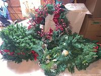 Photo showing pile of green artificial pine garlands and wreaths with red berries, flowers, pine cones, and gift bag in background.
