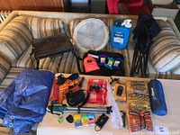 Full lot displayed on couch: tarp, grill top, white plastic fold-out side table, orange kite string kit, folding chairs, grill chips and smoker, and various small camping and kite accessories.