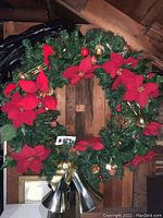Front view of large green wreath decorated with large red poinsettia flowers, gold pinecones, red and gold ornaments, and three gold bells tied with a gold ribbon at the bottom.