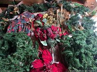 Photo of assorted holiday decor items including green artificial garlands, red poinsettia flowers, gold sticks, and a patterned fabric item arranged together.