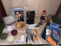 Kitchen items laid out on a table including DeLonghi espresso machine, French press, Tupperware containers, Magic Bullet, cooking utensils, small bowls, knives, gumball machines, and serving tray.