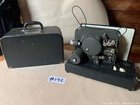 Photo of the Movie-Mite projector with carrying case next to it on floor, showing projector details and condition