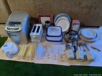 Wide shot showing ice maker, toaster, kettle, dishes, cups, cutlery, plastic containers and straws on table