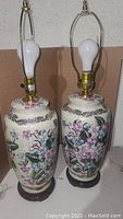 Pair of vintage ceramic table lamps with floral patterns and light green shades, bulbs installed, shown on a surface against a wall.