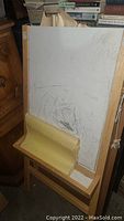 Full view of wooden art easel with white drawing board and brown paper roll holder at the bottom, showing usage marks and sketches on board.