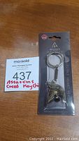 Front view of unopened Assassin's Creed Odyssey Spartan helmet brass keychain in original packaging including MaxSold auction tag.