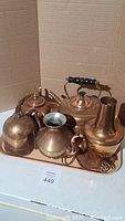 Photo showing six copper/brass colored vintage kitchen items on a tray, including two kettles, two pitchers, a sugar bowl, and a pot.