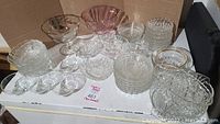 Overview of approximately 40 assorted crystal and glass dishes arranged on a white surface with a cardboard background, showing multiple clear and tinted glass pieces including bowls and plates.