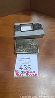 Front view of BC Transit brass belt buckle and original box on wood surface with a lot number card in front.