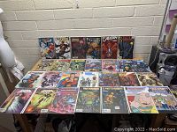 Photo of wide table with 26 assorted comic books arranged in three rows, showing various superhero titles and graphic covers.