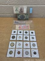Photo showing a 5 Dollar Zimbabwe banknote, a 10 denomination Korean banknote, several other banknotes, and 15 coins placed in individual white holders arranged on a table against a brick wall.