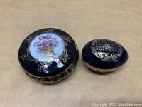 Two Limoges Castel trinket boxes on a wooden surface, showing the top designs and gold detailing.
